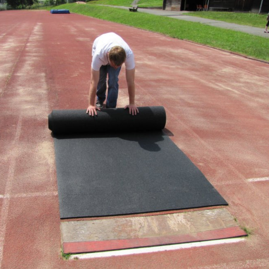 Heavy-duty black rubber roll-out runway for track and field, being unrolled to show the high-traction, slip-resistant surface used for long jump and pole vault.