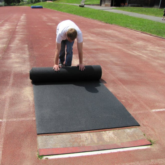 Heavy-duty black rubber roll-out runway for track and field, being unrolled to show the high-traction, slip-resistant surface used for long jump and pole vault.