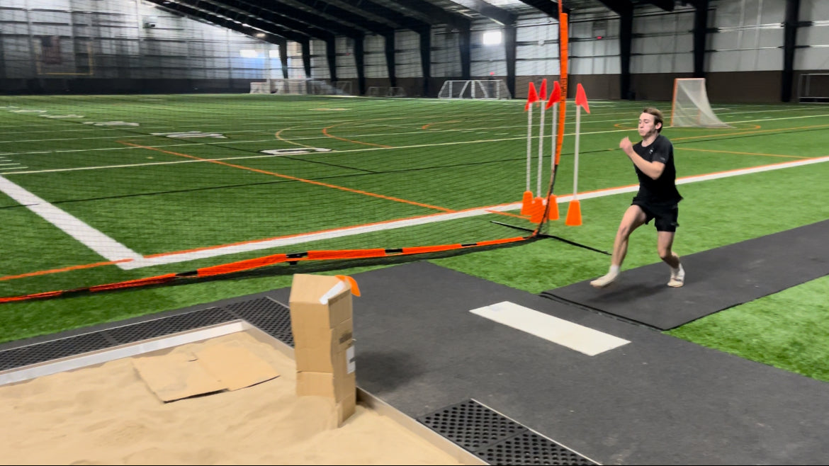 Portable roll-out track runway surface laid out on a practice field, providing a durable and protective rubber lane for triple jump and track events.  Being used inside of a large sports complex.